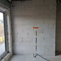 Wewnętrzna ściana z betonu komórkowego w trakcie prac instalacyjnych: widoczne puszki elektryczne, peszel w bruździe i przewody pod sufitem, okno z widokiem na krajobraz.