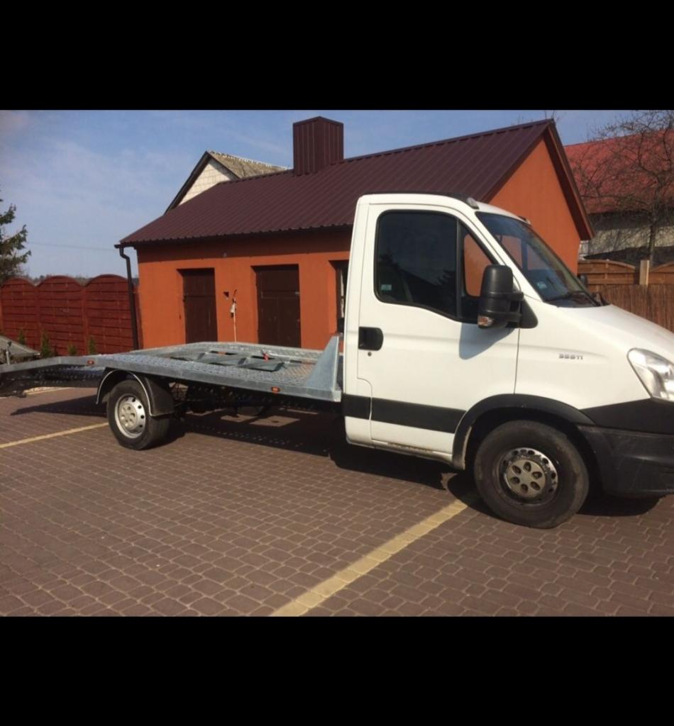 Biała ciężarówka z platformą do transportu pojazdów, zaparkowana na brukowanym placu przed pomarańczowym budynkiem z brązowym dachem.