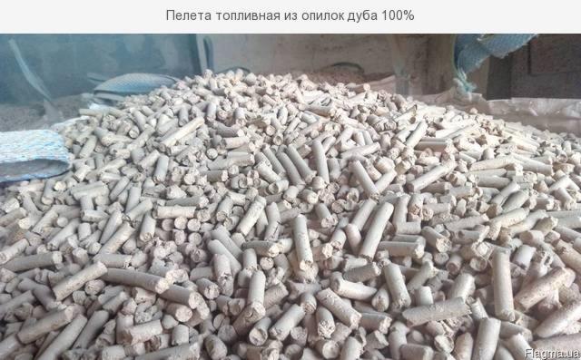 Duża sterta jasnych, cylindrycznych granulek opału w magazynie, widoczne worki w tle.