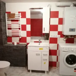 Wykończona łazienka z czerwono-białą szachownicą na ścianach, z białą umywalką z szafką, toaletą, grzejnikiem drabinkowym z ręcznikiem, pralką i piecem gazowym.