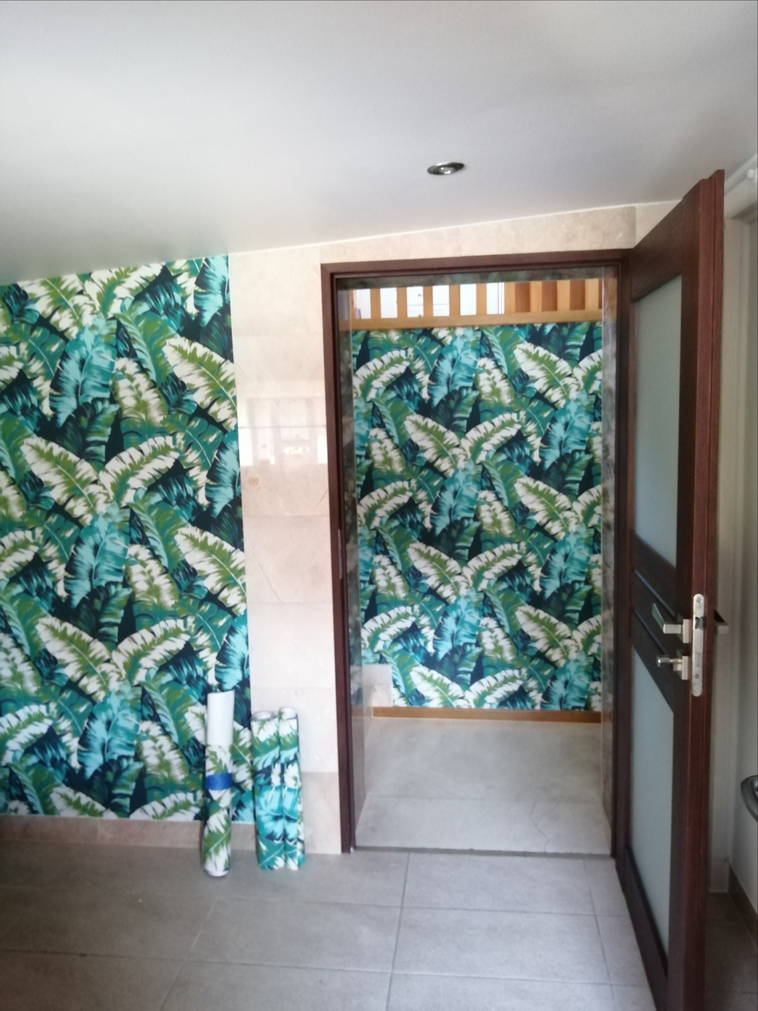 Pokój z wzorzystą tapetą w motywy liści palmowych, z widocznymi rolkami tapety na podłodze, oraz przyległym pokojem również wytapetowanym w ten sam wzór, widocznym przez otwarte drzwi.