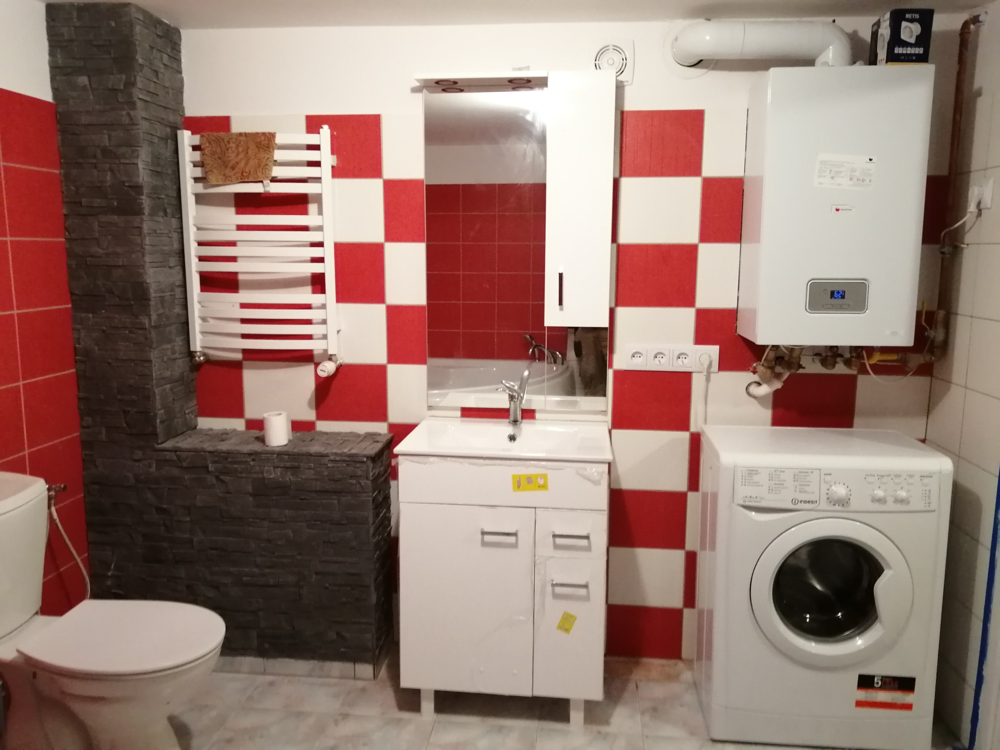 Wykończona łazienka z czerwono-białą szachownicą na ścianach, z białą umywalką z szafką, toaletą, grzejnikiem drabinkowym z ręcznikiem, pralką i piecem gazowym.
