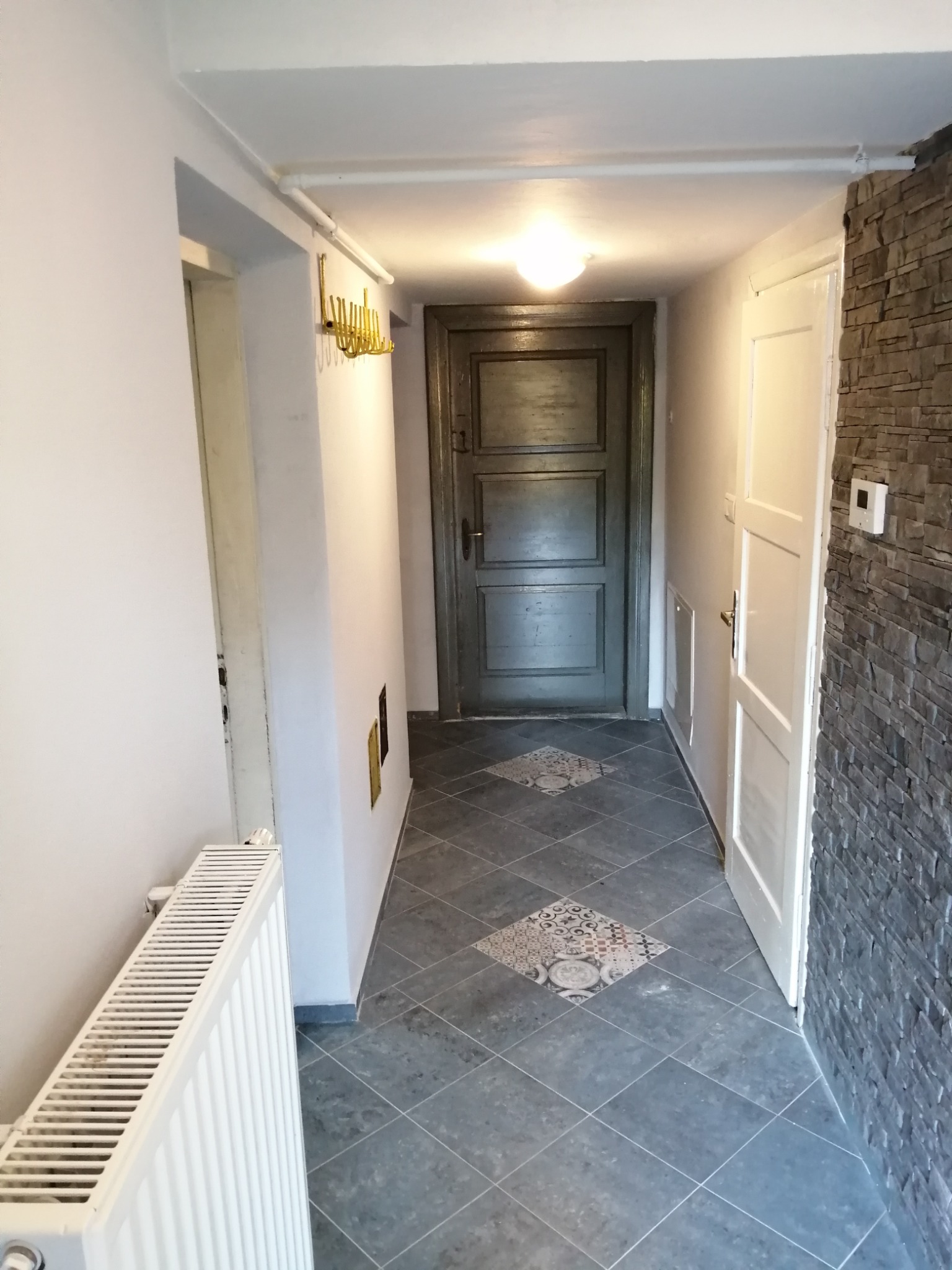 Korytarz po remoncie z szarą płytką podłogową ułożoną w karo, dekoracyjnymi płytkami, białą ścianą, grzejnikiem, kamienną okładziną ściany i ciemnymi drzwiami.