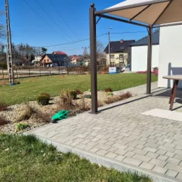 Nowoczesny ogród z trawnikiem, rabatami z kamieni i niskimi krzewami, oraz tarasem z meblami ogrodowymi pod zadaszeniem, w tle domy jednorodzinne.