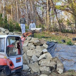 Reality-Designer Jakub Strzałkowski - Prace ziemne w ogrodzie: mini koparka Kubota z widocznym logo firmy MIKOP, dwóch pracowników transportujących kamienie w taczce, czarna agrowłóknina rozłożona na ziemi, drzewa w tle.