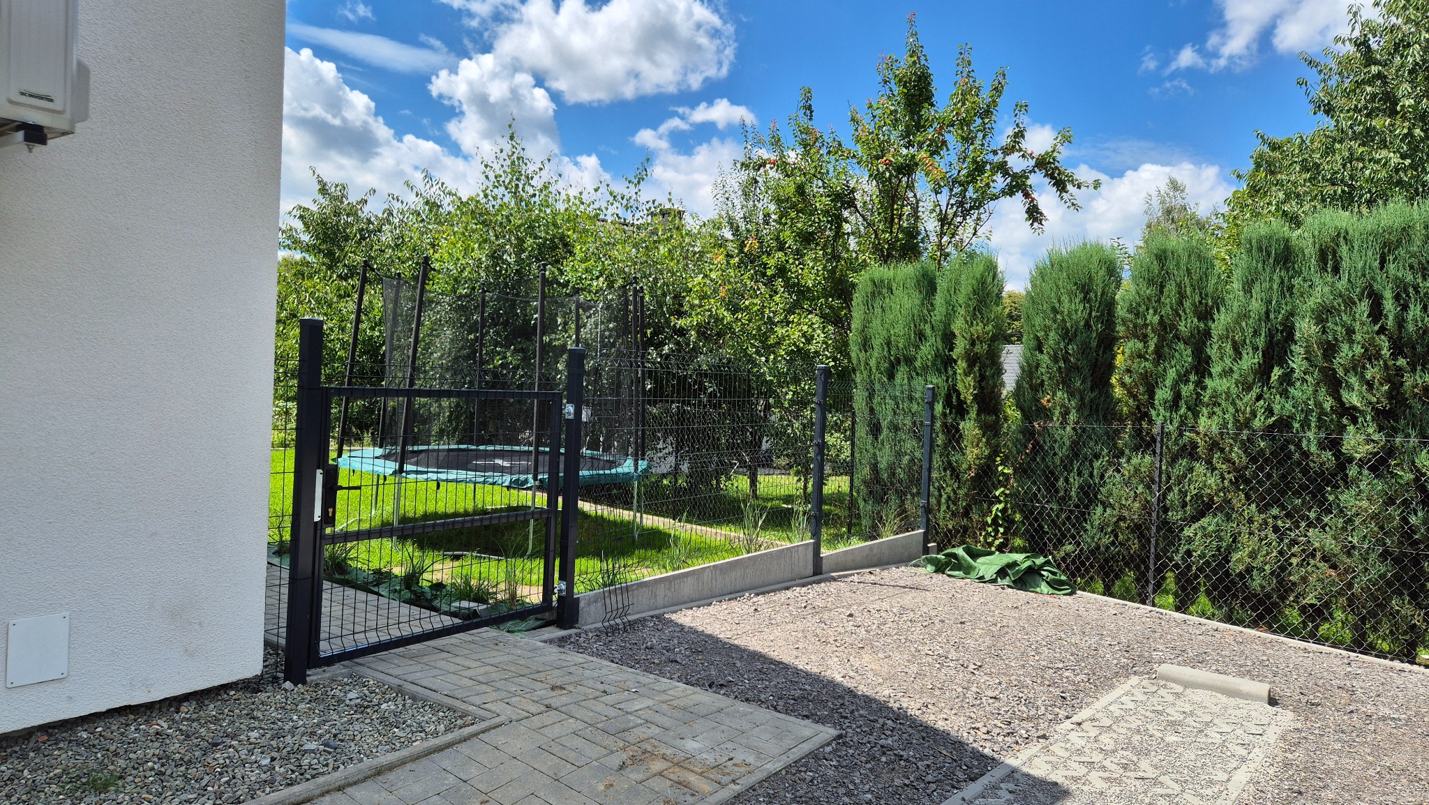 Nowoczesna brama wejściowa z czarnej siatki, prowadząca do ogrodu z zielonym trawnikiem i trampoliną, otoczona żywopłotem z tui i drzewami owocowymi pod błękitnym niebem.