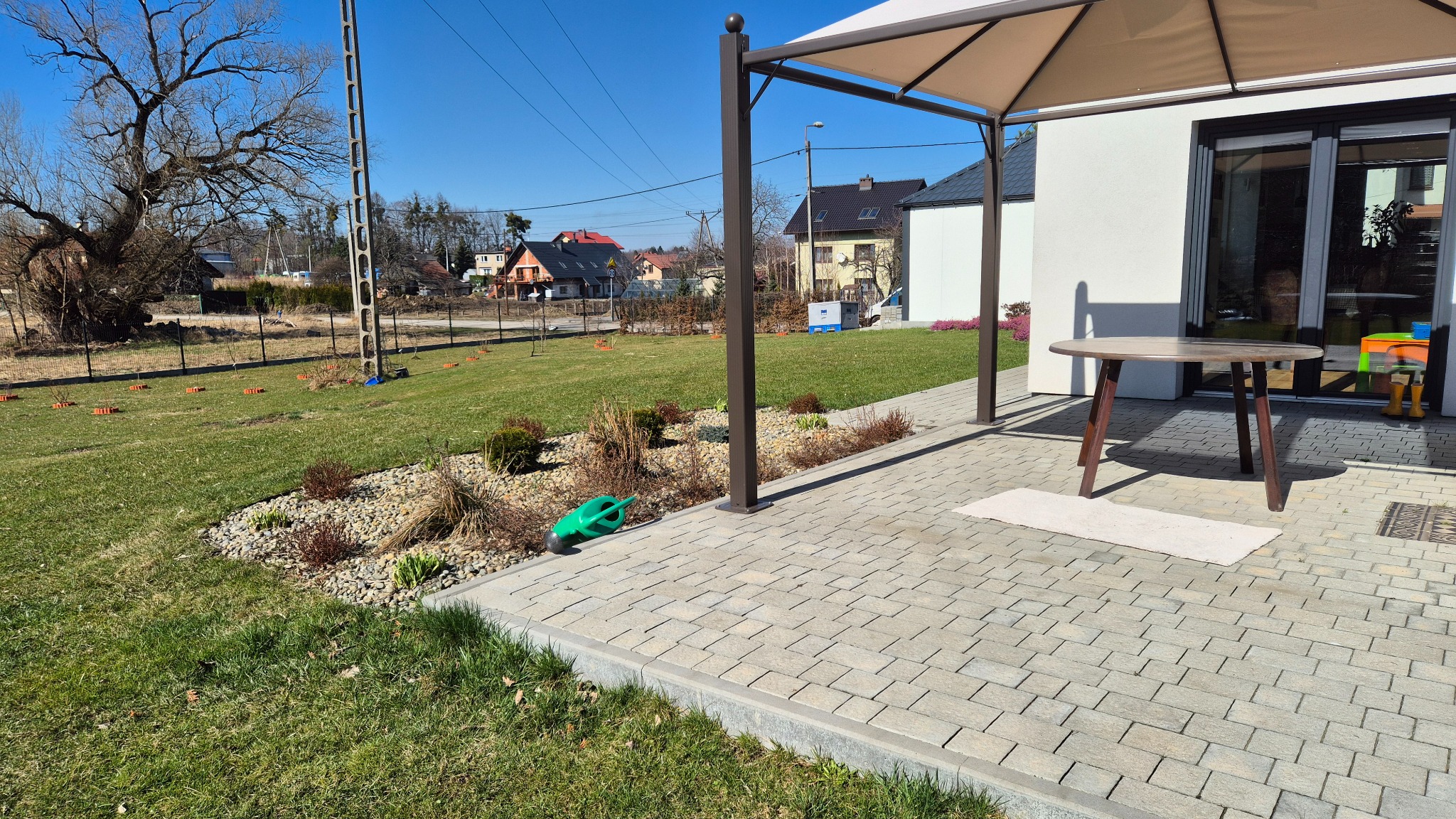 Nowoczesny ogród z trawnikiem, rabatami z kamieni i niskimi krzewami, oraz tarasem z meblami ogrodowymi pod zadaszeniem, w tle domy jednorodzinne.