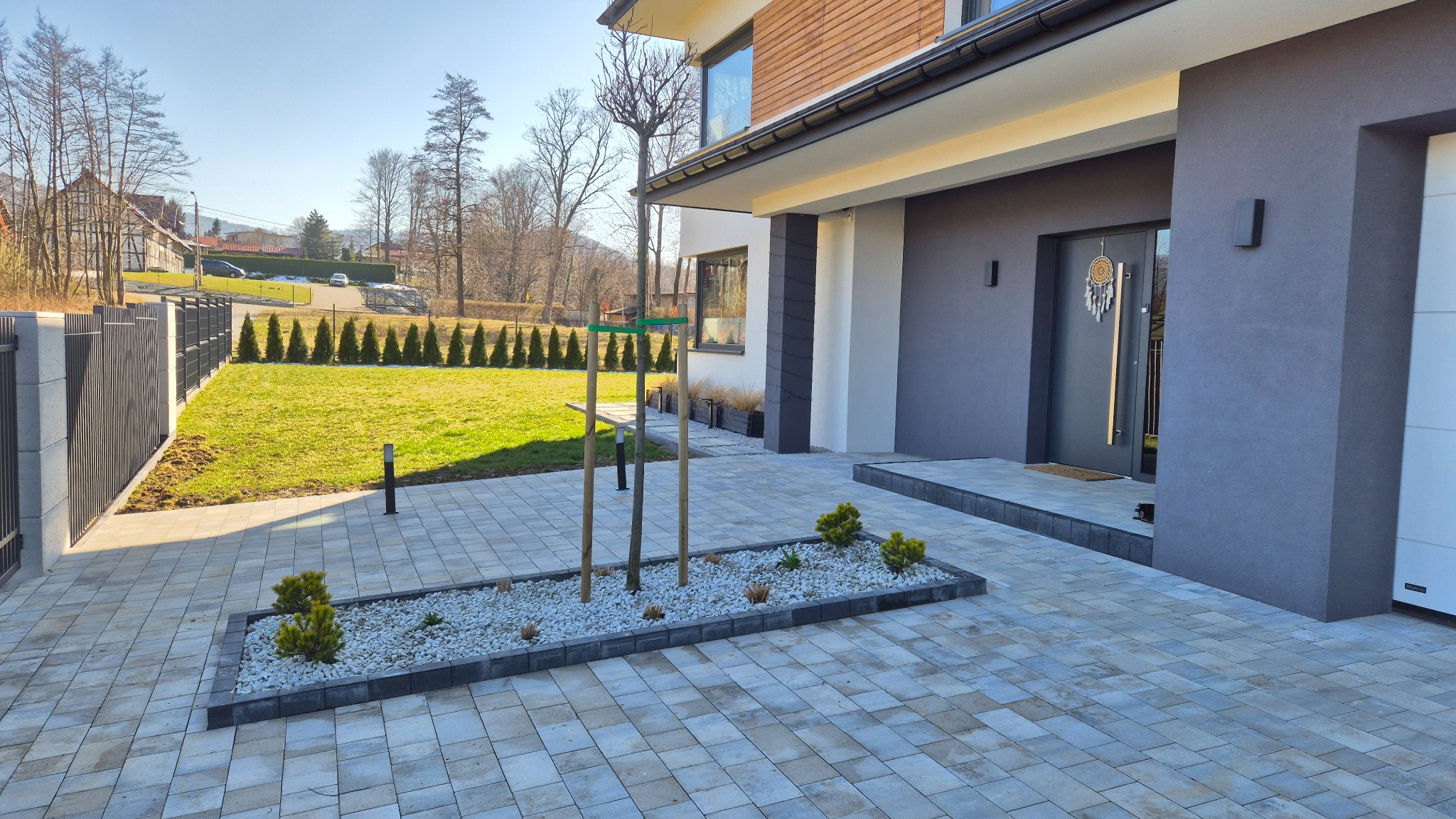 Nowoczesny ogród frontowy z kostki brukowej, rabatą z białym żwirem i młodymi drzewkami, na tle domu z elementami drewna i zadbanego trawnika.