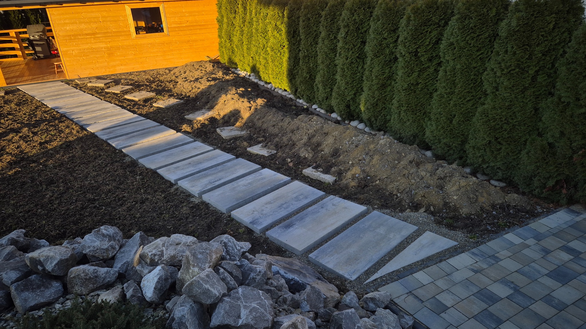 Ścieżka z betonowych płyt prowadząca przez ogród w trakcie tworzenia, z widoczną ziemią, kamieniami i żywopłotem z tui w tle, oświetlona popołudniowym słońcem.