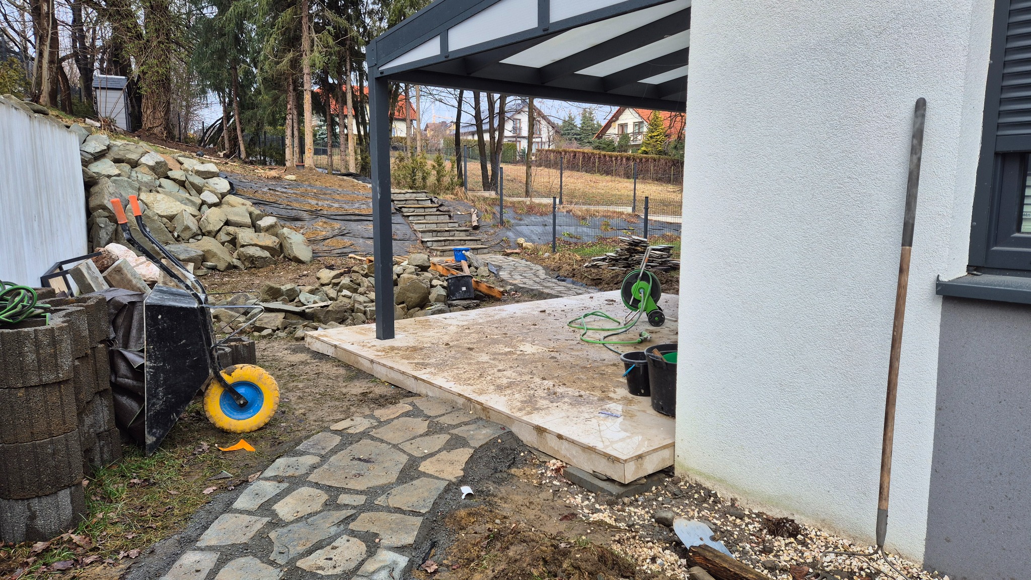Teren w trakcie przygotowania pod budowę tarasu: widoczna podbudowa tarasu, kamienna ścieżka, taczka z materiałami budowlanymi, narzędzia ogrodnicze i kamienny mur oporowy na zboczu.