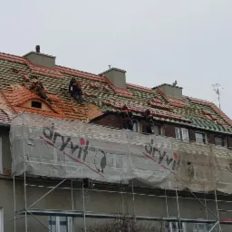 Ekipa montuje czerwoną dachówkę na drewnianej konstrukcji dachu budynku mieszkalnego, widoczne rusztowanie z siatką ochronną z logo Dryvit.
