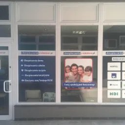 Witryna biura ubezpieczeniowego z naklejkami informacyjnymi o dostępnych ubezpieczeniach, w tym na życie, turystycznych, mieszkaniowych oraz logo różnych towarzystw ubezpieczeniowych.