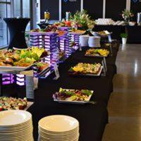 Stół cateringowy z czarnym obrusem, oświetlony fioletowymi lampami LED, zastawiony różnorodnymi przekąskami, sałatkami i deserami. W tle widoczne elementy dekoracyjne i rośliny doniczkowe.