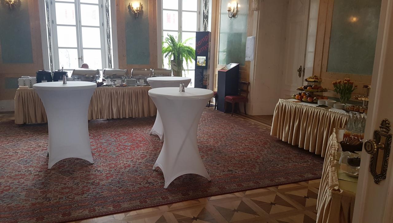 Eleganckie wnętrze z przygotowanym cateringiem: bufet z podgrzewaczami, stół z owocami i deserami, wysokie stoliki koktajlowe w białych pokrowcach, dekoracyjny dywan w klasycznym stylu.