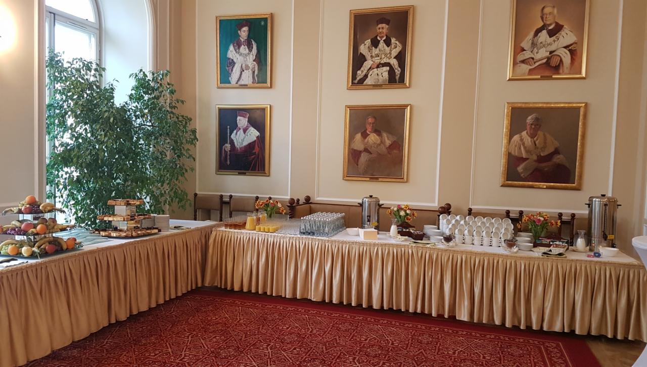 Uroczysty bufet z owocami, ciastami, napojami i kawą, ustawiony w eleganckim wnętrzu z portretami w złotych ramach na ścianach i wzorzystym czerwonym dywanem.