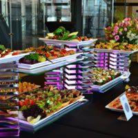 Bogato zastawiony stół z różnorodnymi przekąskami na srebrnych paterach, dekorowany kwiatami, w eleganckim wnętrzu.