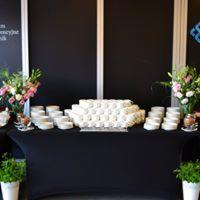 Stół przykryty czarnym obrusem, na nim ułożone piramidalnie białe talerzyki z przekąskami, po bokach kwiaty w wazonach i doniczki z zielonymi roślinami, tło stanowi czarna ściana.