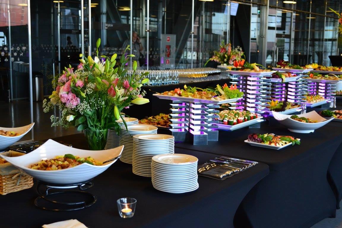 Elegancko zastawiony stół z różnorodnymi potrawami, kwiatami i świecami w szklanym świeczniku, w tle nowoczesna architektura z przeszkleniami.