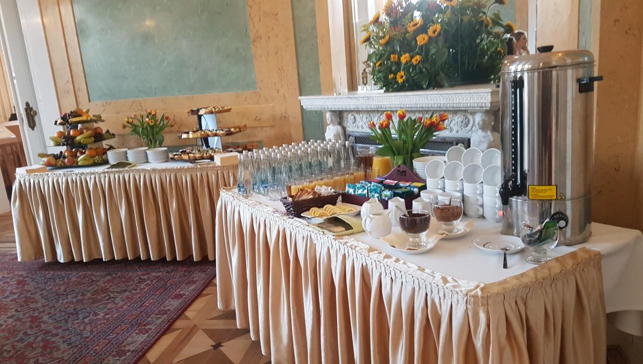 Elegancko zastawiony stół z przekąskami, napojami i ekspresem do kawy, przygotowany na uroczystość w klasycznym wnętrzu.