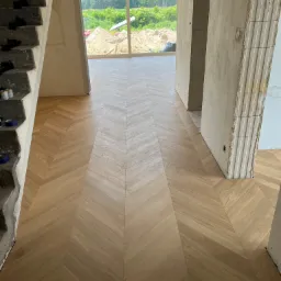 Ułożony parkiet w jodełkę w surowym wnętrzu, widoczny betonowy schody i niedokończone ściany, okno z widokiem na zieleń i piasek.