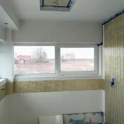 Wnętrze łazienki w trakcie prac wykończeniowych: mozaika na ścianie i suficie, częściowo położone białe płytki, widoczne okno z widokiem na krajobraz.