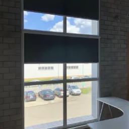 Dwa okna z zasuniętymi roletami wewnętrznymi w pomieszczeniu biurowym z widokiem na parking.