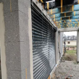 Szare żaluzje fasadowe zamontowane na surowym budynku z bloczków betonowych, widoczne narzędzia budowlane i materiały izolacyjne.