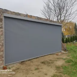 Szara, zwinięta przesłona na tarasie z kamienną ścianą w tle, widok na ogród z tujami i nagie drzewa w pochmurny dzień.