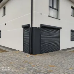 Nowoczesny dom z szarymi roletami zewnętrznymi na oknach i drzwiach, widok z poziomu kostki brukowej na podjeździe.