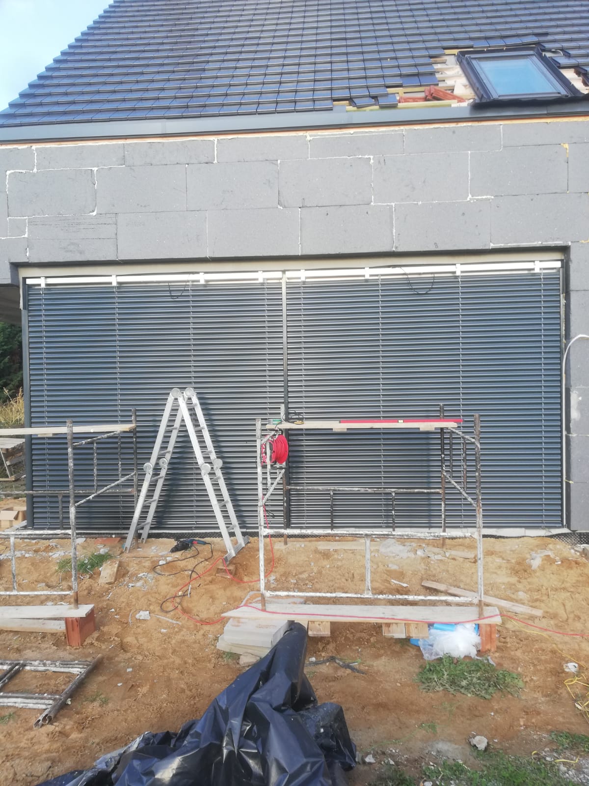 Montaż żaluzji fasadowych na elewacji budynku z wykorzystaniem rusztowania i drabiny, widoczne narzędzia i materiały budowlane.