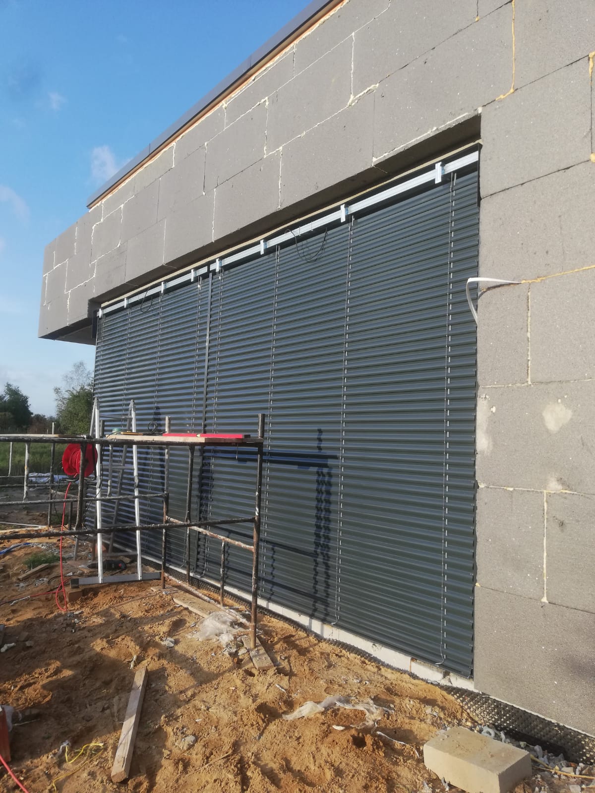 Montaż ciemnoszarych, poziomych osłon okiennych na surowej elewacji budynku, widoczne rusztowanie i narzędzia.