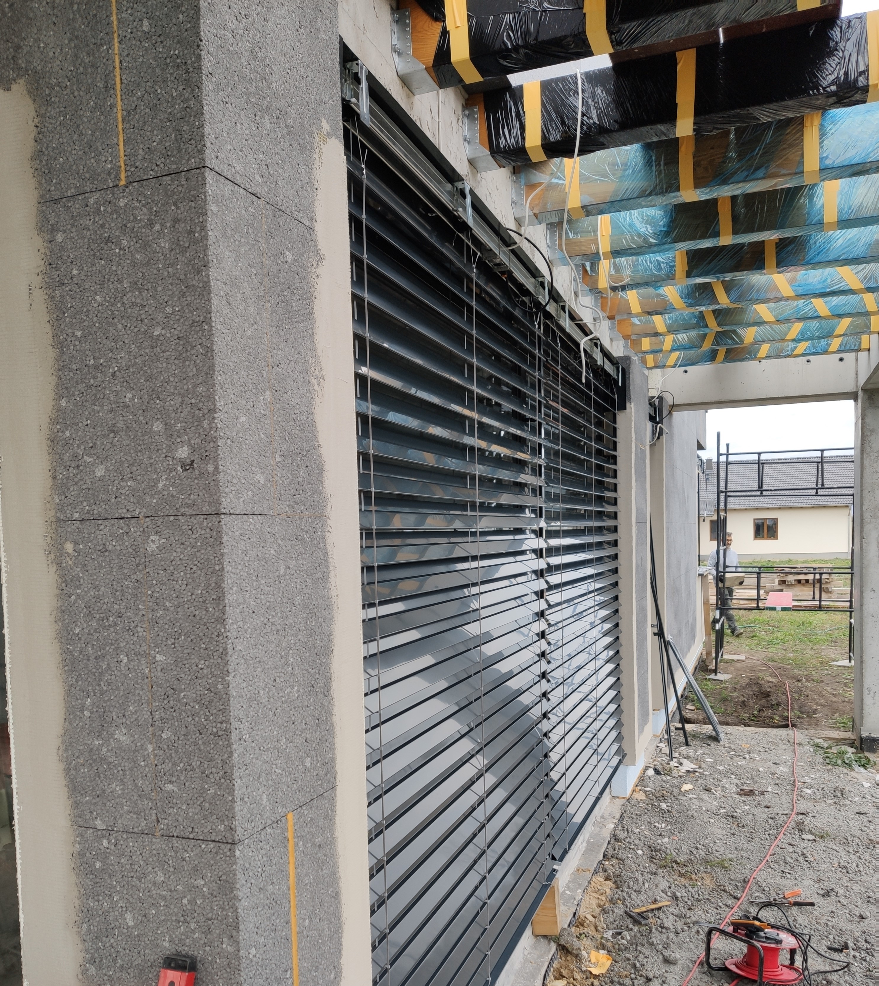 Szare żaluzje fasadowe zamontowane na surowym budynku z bloczków betonowych, widoczne narzędzia budowlane i materiały izolacyjne.