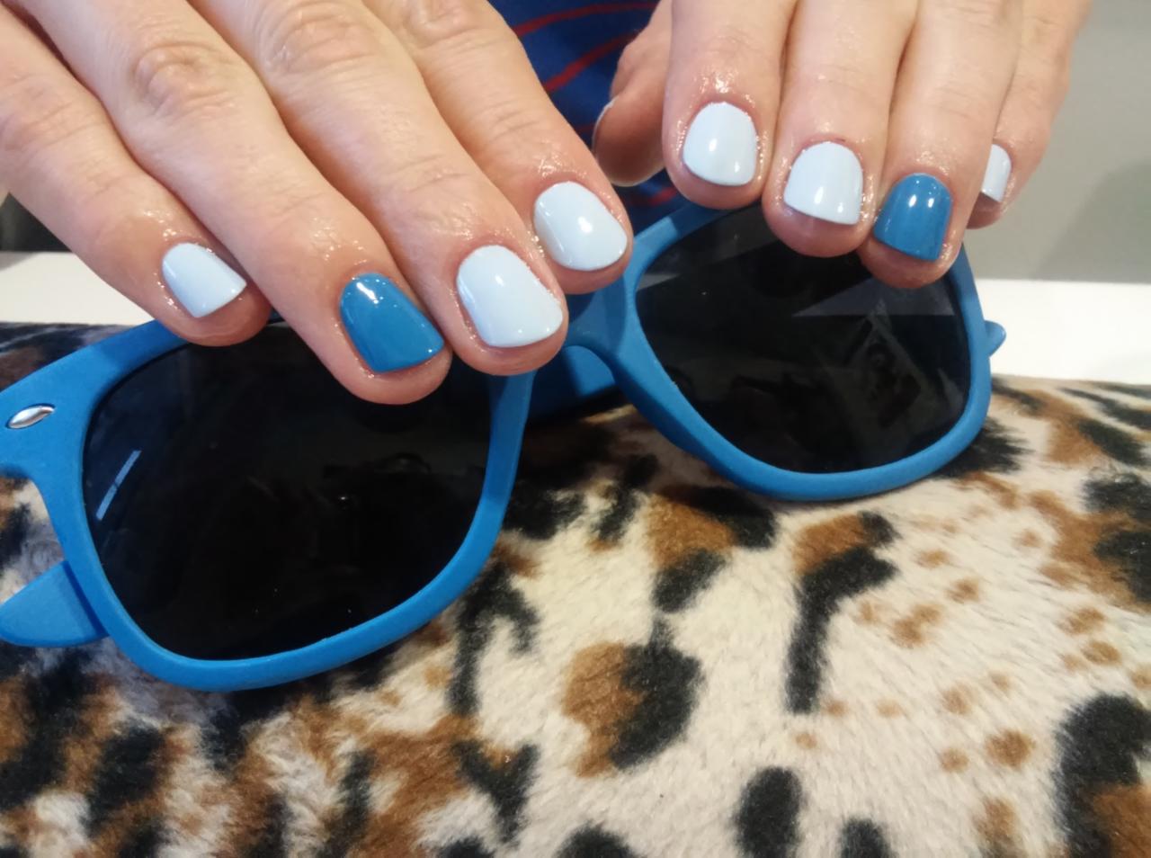 Świeżo pomalowane paznokcie w odcieniach błękitu i baby blue, dłonie na niebieskich okularach przeciwsłonecznych z wzorem panterki w tle.