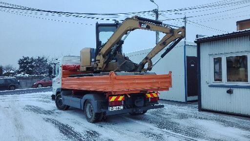 Pomarańczowa wywrotka z żółtą mini-koparką CAT na skrzyni ładunkowej, stojąca na zaśnieżonym placu przy budynku.