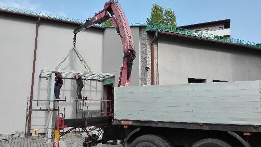 Montaż konstrukcji stalowej zadaszenia nad wejściem do budynku, z użyciem dźwigu i rusztowania, pracownicy w trakcie mocowania.