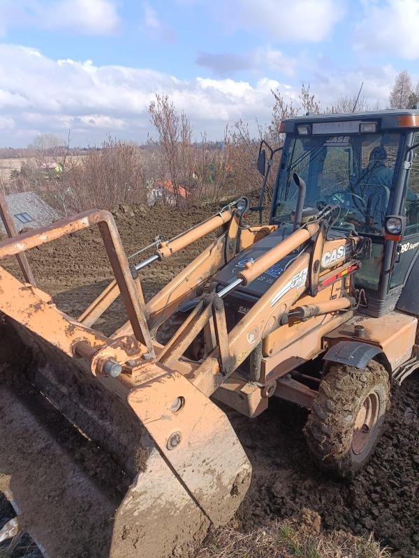 Pomarańczowa koparko-ładowarka CASE 580 Super LE z łyżką zanurzoną w ziemi, widok z bliska na tle krajobrazu z drzewami i domami w oddali, błękitne niebo z chmurami.