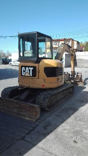 Żółta mini koparka CAT na gąsienicach, zaparkowana na betonowym placu, z widocznymi śladami użytkowania i zarysowaniami na karoserii.