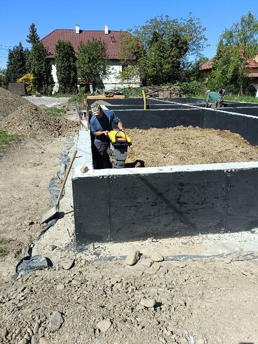 Prace ziemne przy fundamentach domu jednorodzinnego: ubijanie gruntu ubijarką wibracyjną, widoczne czarne izolowane ściany fundamentowe, w tle dom i drzewa.