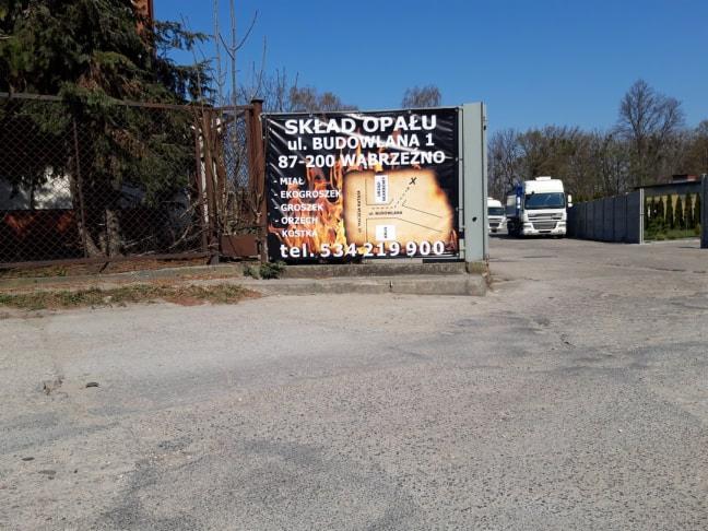 Tablica reklamowa z nazwą i adresem firmy oferującej różne rodzaje opału, widoczna z drogi dojazdowej, z dwoma białymi ciężarówkami w tle.