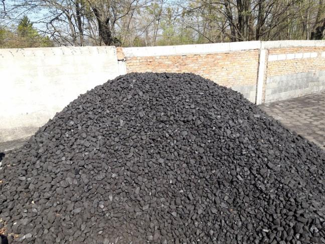 Góra czarnego opału ułożona na placu, w tle fragment muru z betonu i cegły.