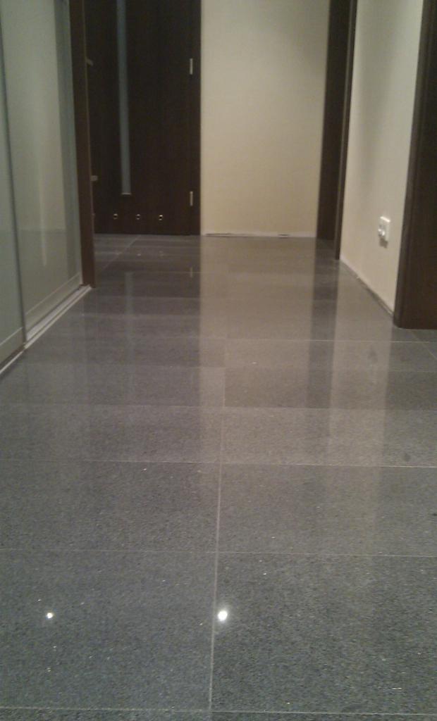 Połyskująca, ciemnoszara posadzka z płytek w korytarzu, widoczne drzwi w ciemnej ościeżnicy i fragment ściany z gniazdkiem.