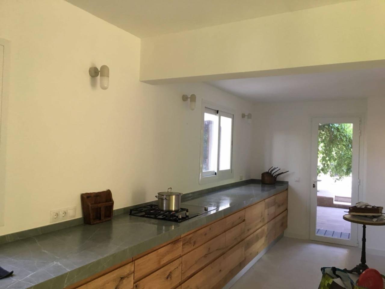 Jasna kuchnia z długim blatem z szarego marmuru, drewnianymi szafkami z wyraźnym usłojeniem i płytą grzewczą z garnkiem. Widoczne gniazdka elektryczne, drewniany organizer, okno i przeszklone drzwi...