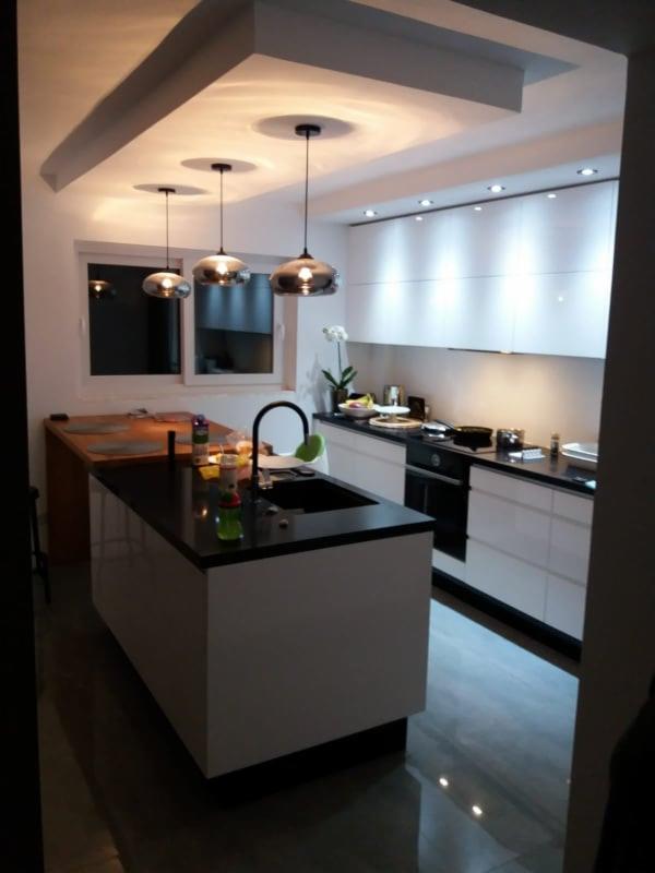 Nowoczesna kuchnia z wyspą i błyszczącymi białymi szafkami, oświetlona trzema szklanymi lampami wiszącymi nad blatem z czarnego kamienia, z widocznym fragmentem drewnianego blatu w tle.