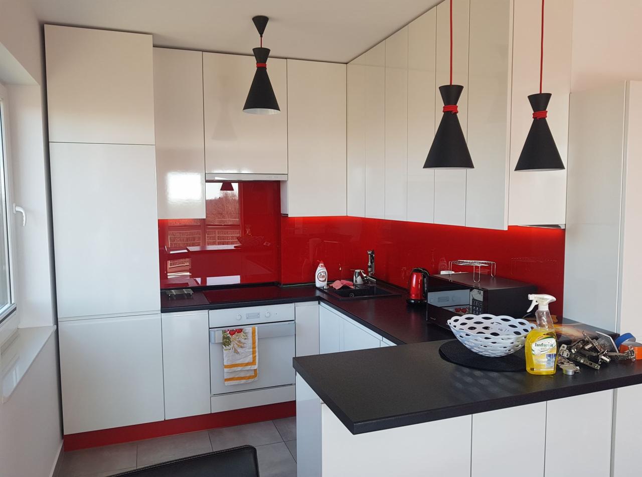 Biała kuchnia z czerwonym panelem szklanym między szafkami, czarnym blatem i trzema czarnymi lampami stożkowymi z czerwonymi akcentami. Na blacie kuchennym widoczny płyn do mycia naczyń i akcesoria...