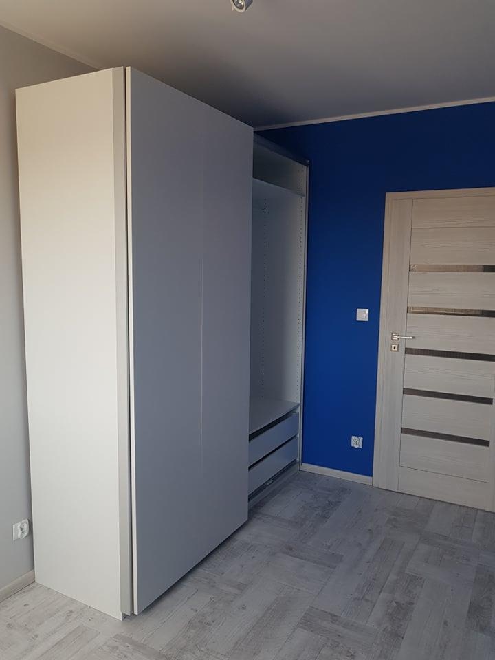 Wysoka, dwukolorowa szafa z szarymi frontami i białym bokiem, z widocznymi szufladami wewnątrz, na tle niebieskiej ściany i jasnoszarej podłogi w jodełkę.