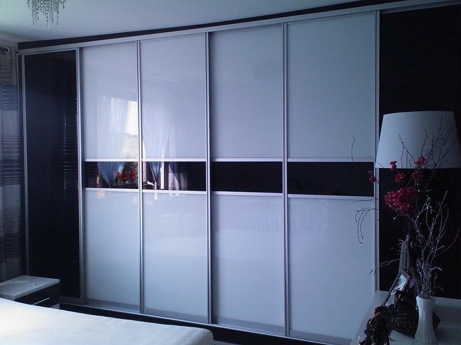 Duża szafa przesuwna z białymi i czarnymi panelami, srebrne obramowanie, widoczna lampka nocna i dekoracje na szafce nocnej.