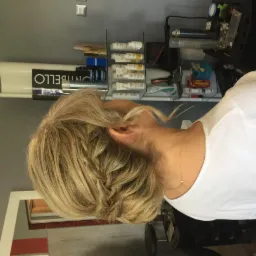 Blond włosy upięte w warkocz, widok z boku, w tle półka z kosmetykami i akcesoriami.