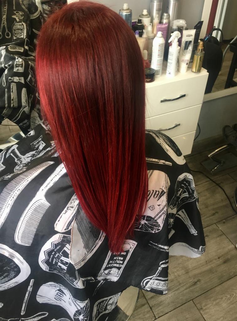 Świeżo ufarbowane, intensywnie czerwone, proste włosy, widoczne z tyłu, na tle białej komody z kosmetykami i peleryny ochronnej z motywem fryzjerskim.