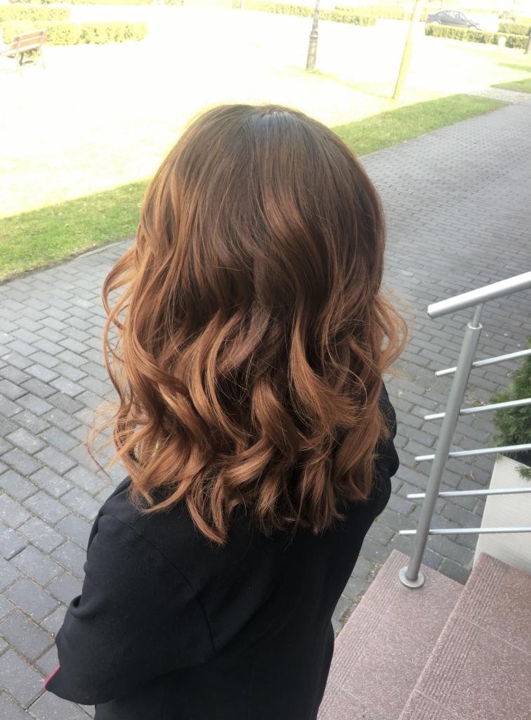 Kobieta z brązowymi, falowanymi włosami z refleksami, sfotografowana z tyłu na zewnątrz, w czarnej marynarce, stojąca na kostce brukowej obok schodów z metalową barierką.