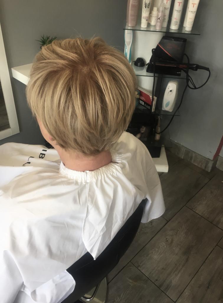 Krótkie, blond włosy po strzyżeniu, widok z tyłu, na głowie peleryna ochronna, w tle półki z kosmetykami i sprzętem.